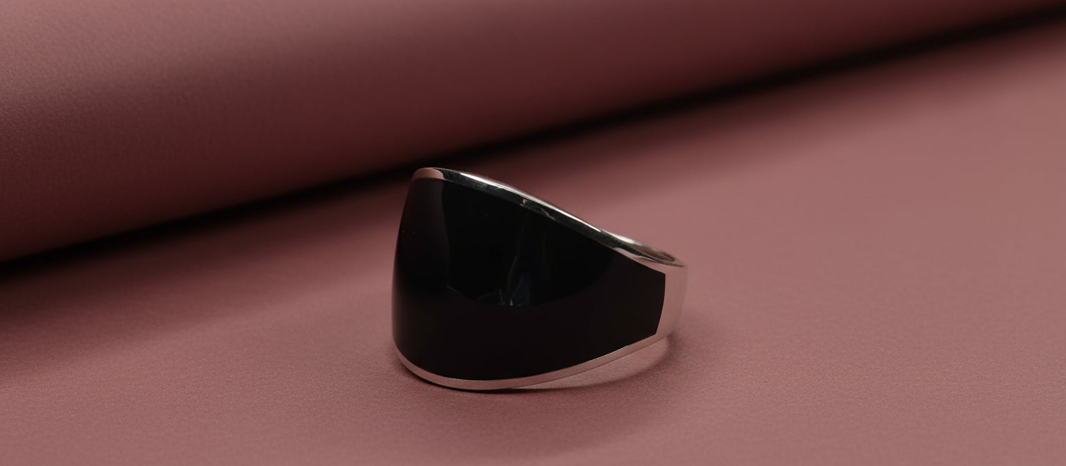 The Onyx Shield Ring from Narayan Jewellers, a wide sterling silver band with a large, curved black enamel face, on a dusty rose background.